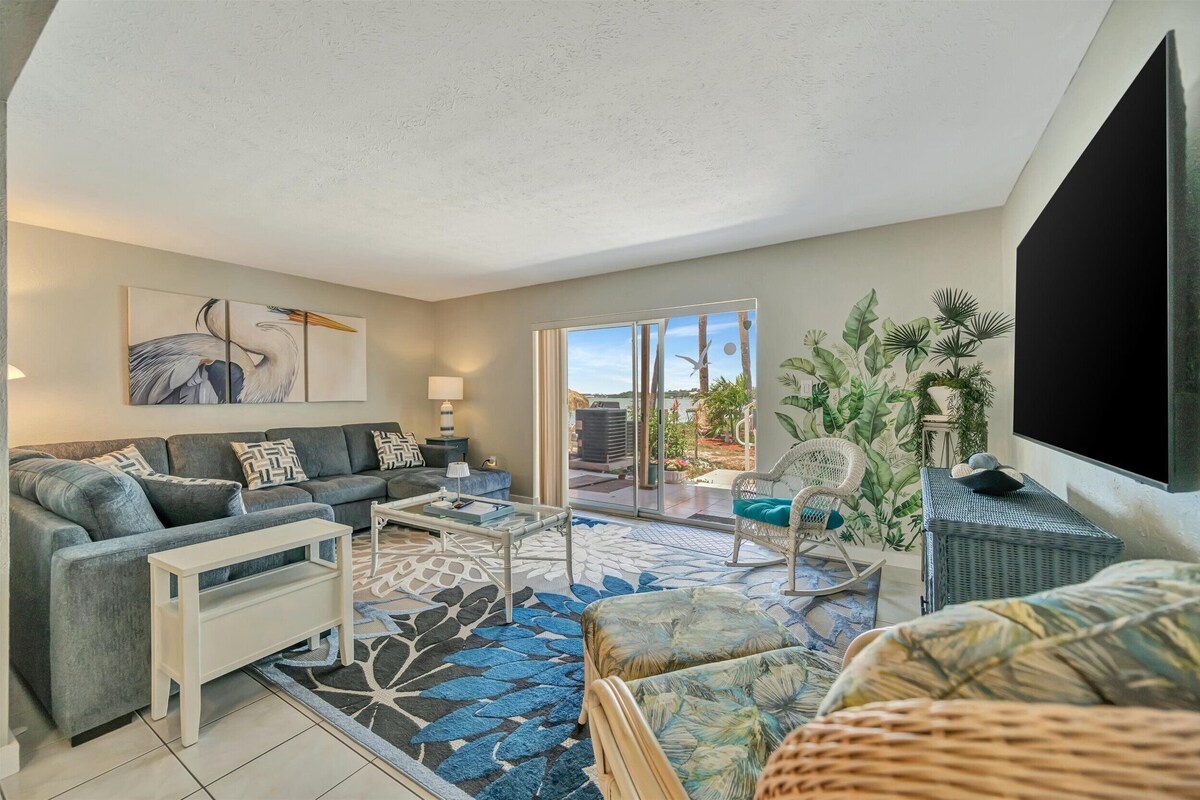 A living room with a sofa, coffee table, and a large TV. There is a balcony with plants and a view of the outdoors.