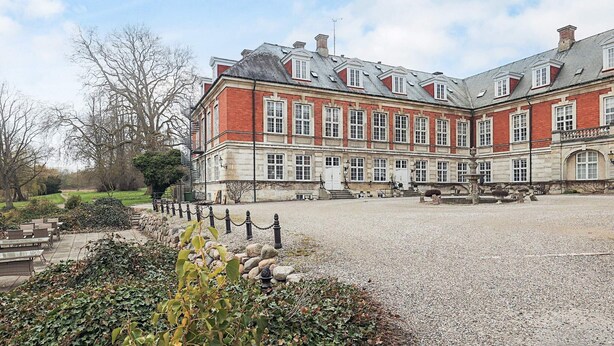 A large, historic building with a symmetrical facade, multiple windows, and a gravel courtyard.