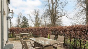 An outdoor patio with a table and chairs, surrounded by a hedge and trees.