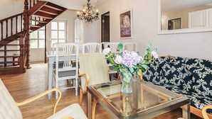 A spacious living area with a wooden staircase, a glass coffee table, white chairs, a sofa with a patterned cover, and a chandelier.