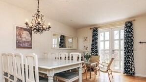 A dining room with a white table and chairs, a chandelier, and a large window with curtains.