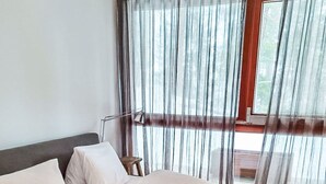 Ein ordentlich bezogenes Bett mit Kissen und Decke, ein Nachttisch mit Lampe und ein Fenster mit Vorhängen.