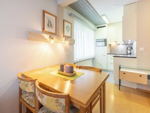A dining area with a wooden table and chairs, a small kitchen with a coffee maker, and a window with curtains. - Val Signina 7-1 by Interhome (Laax)