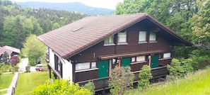 Ein zweistöckiges Holzhaus mit geneigtem Dach, grünen Türen und Fenstern mit Vorhängen.