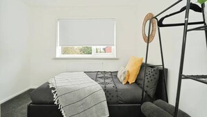 Une pièce avec un canapé noir, une couverture grise, un coussin jaune, un fauteuil suspendu noir et une fenêtre avec un store blanc.