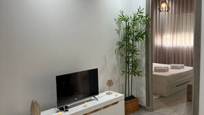 En moderne stue med et fladskærms-tv på et hvidt skænk, en stor stueplante og en seng med udsigt gennem et vindue med gardiner.