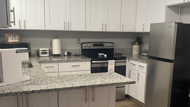 Una cocina con armarios blancos, encimera de granito, refrigerador de acero inoxidable y mesa de comedor de madera.