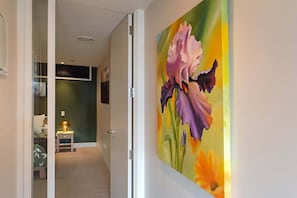 A hallway with a door leading to a room, a painting of flowers on the wall, and a lamp on a nightstand. - Moana Sands - Beachside Apartment (Nelson)