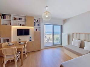Ein modernes Wohnzimmer mit Essbereich, einem an der Wand befestigten Fernseher, einem großen Fenster mit Meerblick und einer gemütlichen Sitzecke mit Sofa und Kissen.