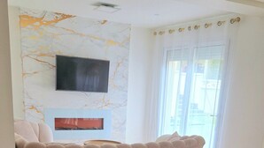 A living room with a sofa, a flat-screen TV mounted on the wall, a fireplace, and a large window with curtains.