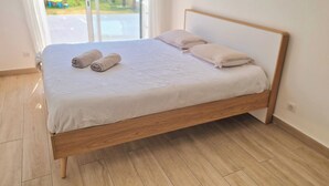A neatly made bed with a wooden frame and white bedding, a wooden floor, and a view of a playground through a glass door.