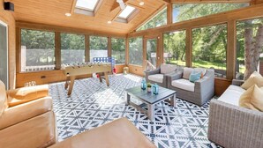 A sunlit room with a foosball table, wicker furniture, and large windows. - Linn Lodge | Fire Pit, 5+ Night Stays Include Golf Cart | Sleeps 10 (Lake Geneva)