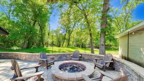 A circular fire pit surrounded by Adirondack chairs, set in a brick patio with a view of a grassy yard and trees. - Linn Lodge | Fire Pit, 5+ Night Stays Include Golf Cart | Sleeps 10 (Lake Geneva)