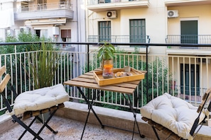 Ein Balkon mit Holztisch, zwei gepolsterten Stühlen, einem Blumentopf und einer Ablage mit Speisen und Getränken.