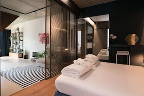 Une chambre dâhĂŽtel moderne dotĂ©e dâun grand lit, dâune cloison en verre, dâun coin salon avec un canapĂ© et dâune petite table.