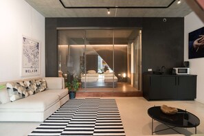 Een moderne woonkamer met een bank, een zwart-wit kleed, een glazen deur naar een slaapkamer en een zwart kastje.
