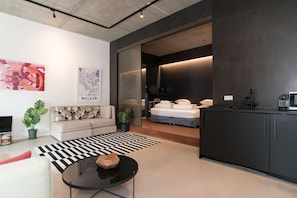 Un salon moderne avec un canapĂ©, une table basse et des Ćuvres dâart. On aperçoit un lit Ă lâarriĂšre-plan, avec un mur sombre et une source de lumiĂšre.
