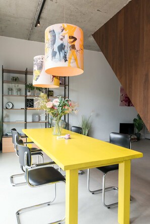 Een gele eettafel met stoelen, een vaas met bloemen en een grote lamp met een poster.