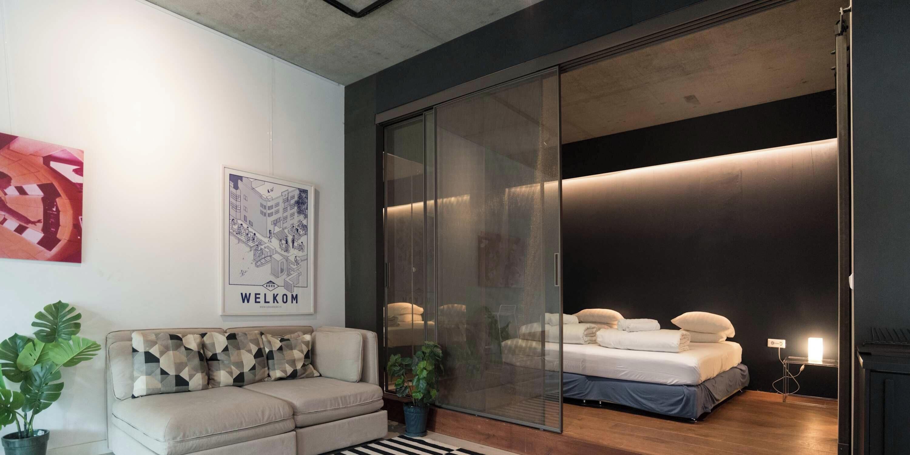 Un dormitorio moderno con puerta de cristal corredera que da a un balcón, un sofá con cojines estampados y un cartel de bienvenida enmarcado.