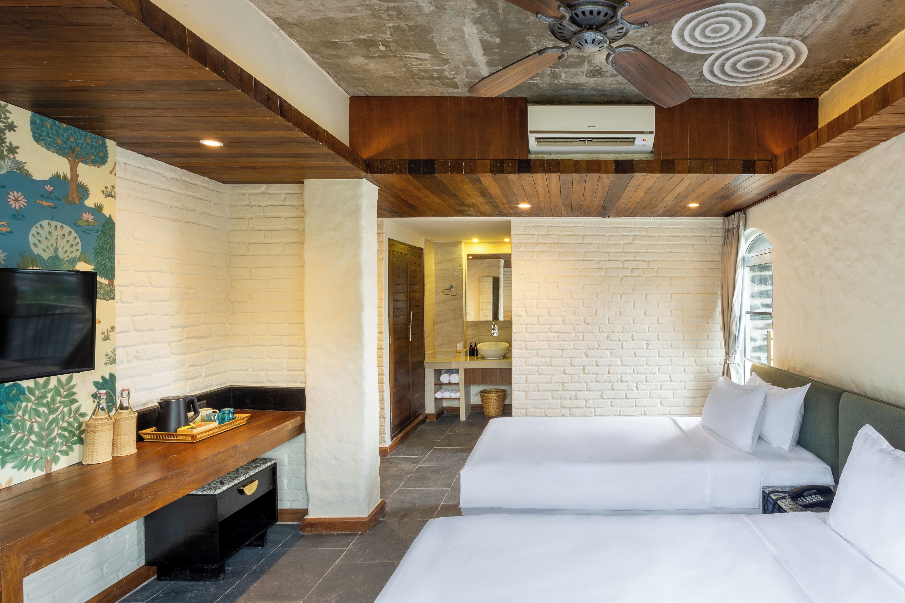 A hotel room with a wooden ceiling, a flat-screen TV, a brick wall, a bed, and a bathroom visible through an open door.