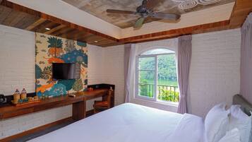 A hotel room with a bed, a desk with a TV, a window with a view, and a ceiling fan.