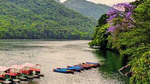 A calm lake with boats docked on the shore, surrounded by lush green hills. - Nomad Pokhara Hotel  (Pokhara)
