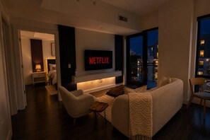 A modern living room with a large TV displaying 'NETFLIX,' a sofa, chairs, a coffee table, and a view of the city at night. - Buckhead Atlanta - Modern Luxury 1BR - Free Parking (Atlanta)