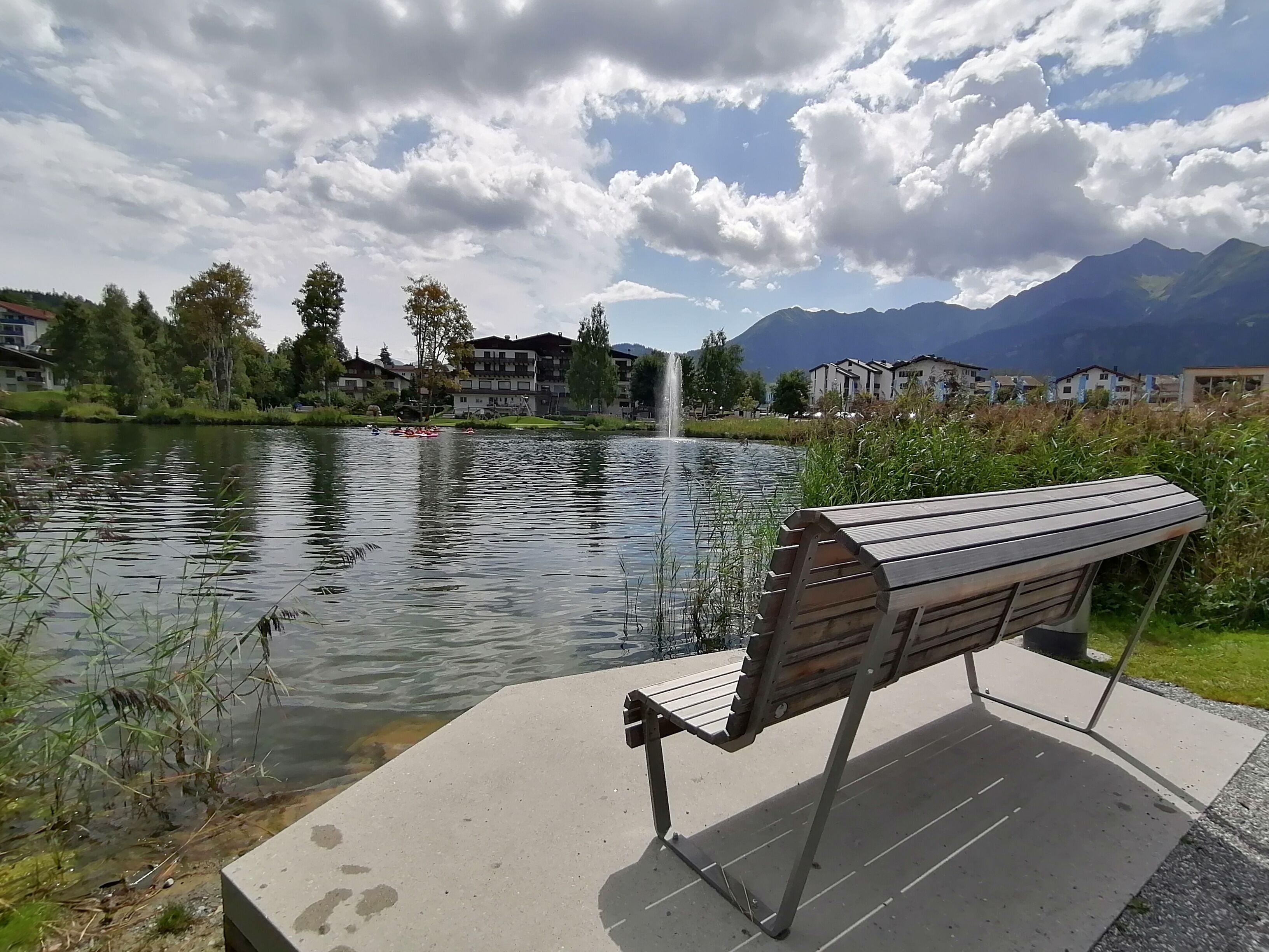 Eine Bank am See mit einem Brunnen, umgeben von Grünflächen und Gebäuden.