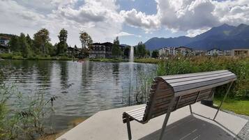 Eine Bank am See mit einem Brunnen, umgeben von Grünflächen und Gebäuden.
