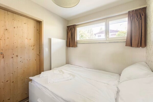 Une chambre avec une porte en bois, une fenêtre avec des rideaux, un lit avec une literie blanche et un radiateur.