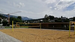 A sandy volleyball court with a wooden deck, surrounded by buildings and mountains.