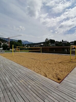 A sandy volleyball court with a wooden deck, surrounded by buildings and mountains.