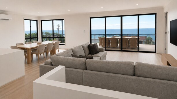 A modern living room with a large gray sofa, a dining area with a wooden table and chairs, and large sliding glass doors offering a view of the ocean. - Wake to Ocean Views & Wildlife at Ohana Ocean Breeze (Penneshaw)