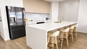 A modern kitchen with a central island, bar stools, a stainless steel refrigerator, and light-colored cabinets. - Wake to Ocean Views & Wildlife at Ohana Ocean Breeze (Penneshaw)