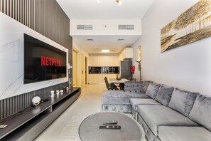 Ein modernes Wohnzimmer mit einem großen Flachbildfernseher, einem grauen Ecksofa, einem runden Couchtisch und einer Küche im Hintergrund.
