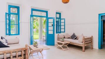 A room with a sofa, a coffee table, and a chair. There are three blue-framed windows and a door.