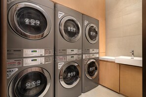 A row of six LG commercial washers with digital displays, a sink, and wooden cabinetry in a laundry room. - Hilton Garden Inn Shanghai Wujiaochang (Shanghai)