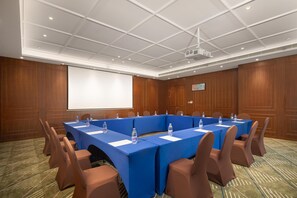 Sala de conferências com uma mesa comprida, cadeiras marrons e uma tela de projeção.