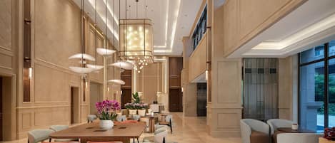 A spacious hotel lobby with a large dining table, comfortable seating, and a modern chandelier.