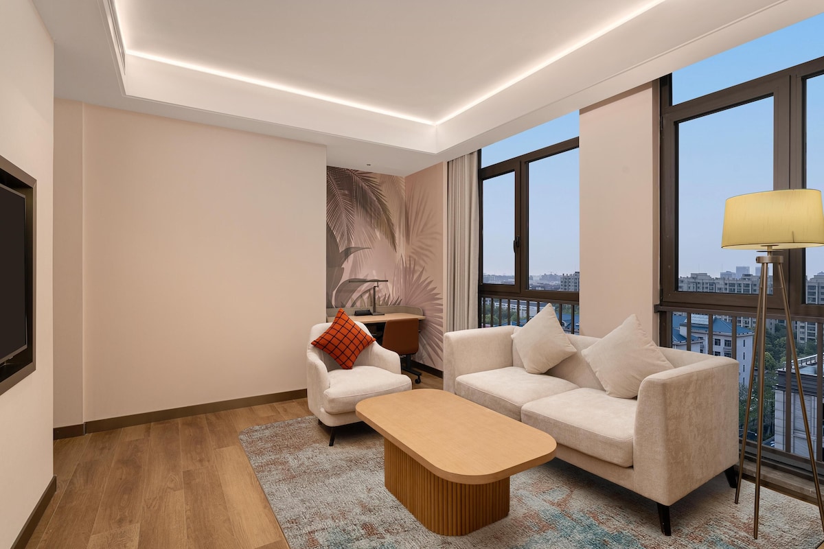 Et moderne hotelværelse med et stort vindue, en sofa, et sofabord og et skrivebord med en lampe.