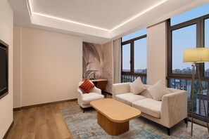 Uma sala de estar moderna com sofá, poltrona e mesa de centro, grandes janelas com vista para a cidade e uma TV de tela plana.