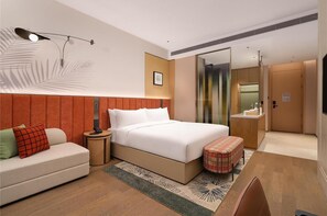 Et moderne hotelværelse med seng, natbord, sofa, lille skærmer, et spejl og et badeværelse med vask og brusebad.