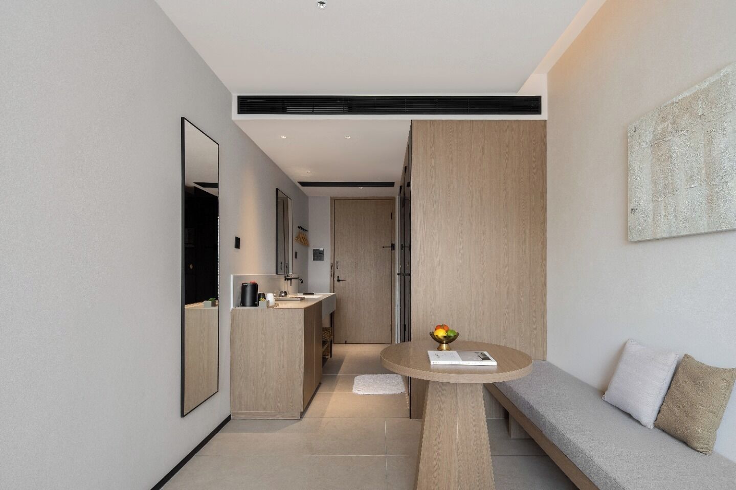 A modern hotel room with a minimalist design, featuring a large mirror, a round table with a book and fruit, a sofa with cushions, and a wooden accent wall.