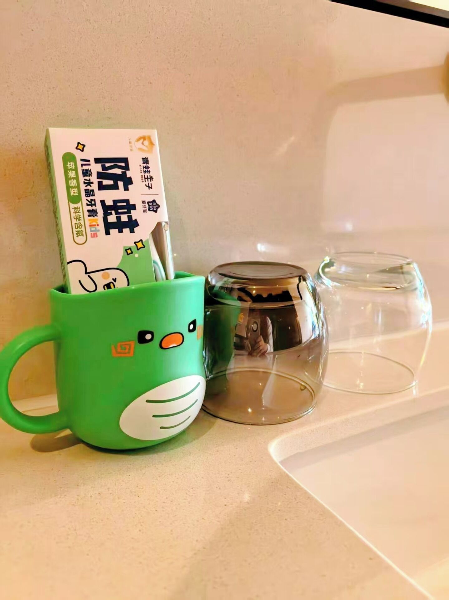 Una taza verde con cara de caricatura, una taza de vidrio transparente y una caja de fideos instantáneos sobre una encimera.