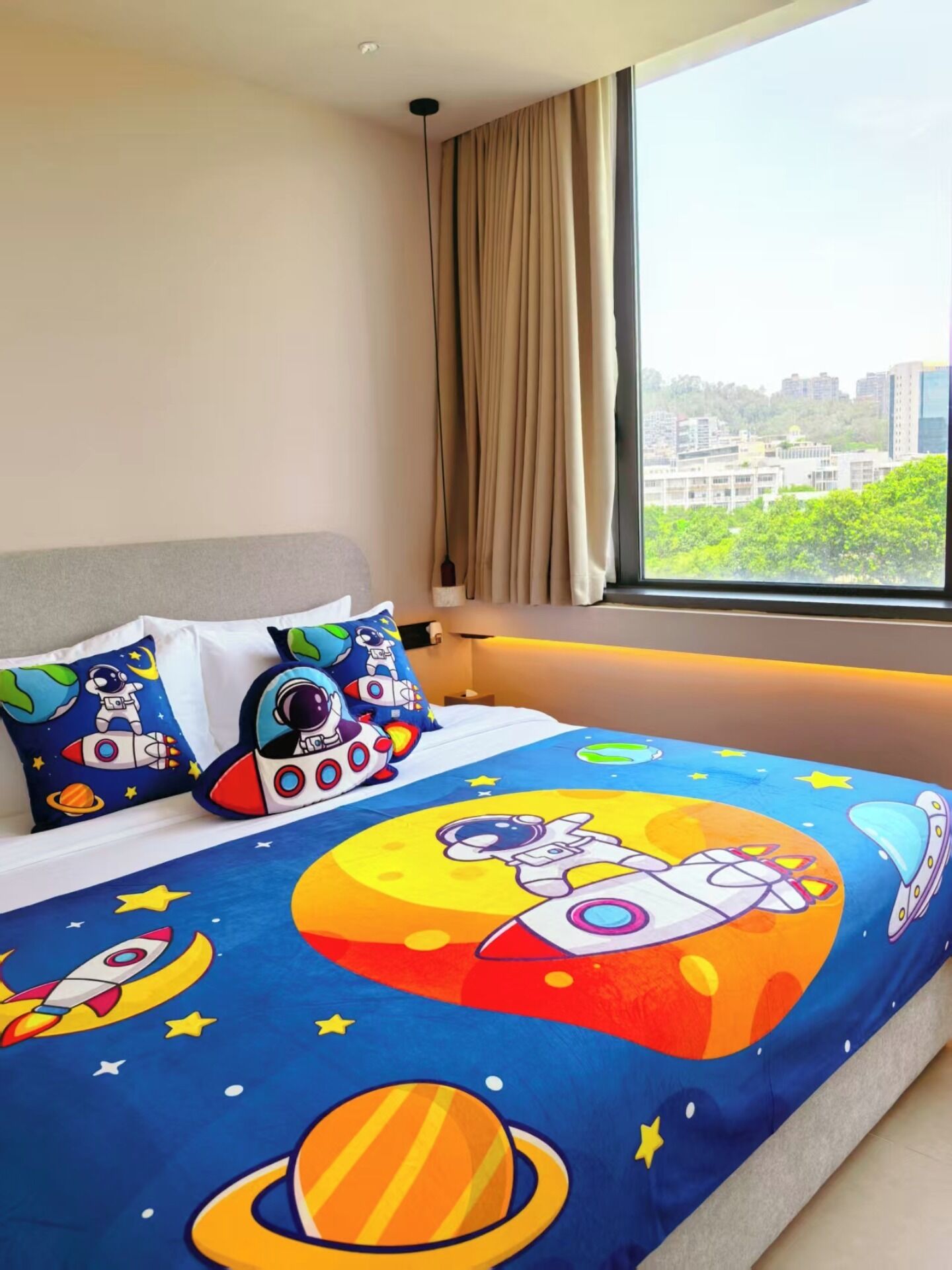 A bedroom with a bed featuring a space-themed bedspread, a view of a cityscape through a large window, and a curtain on the window.
