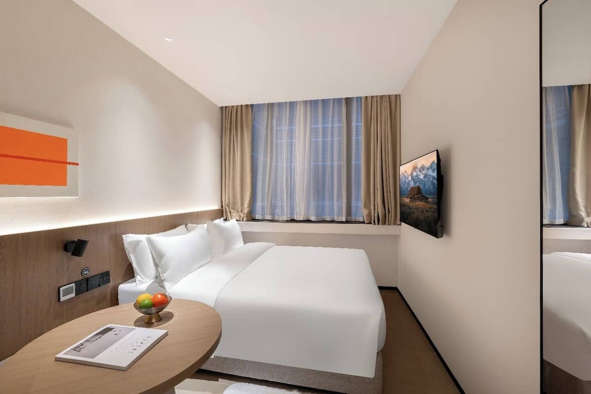Una habitación de hotel con una cama, una mesita de noche con un libro y fruta, una ventana con cortinas y un cuadro en la pared.