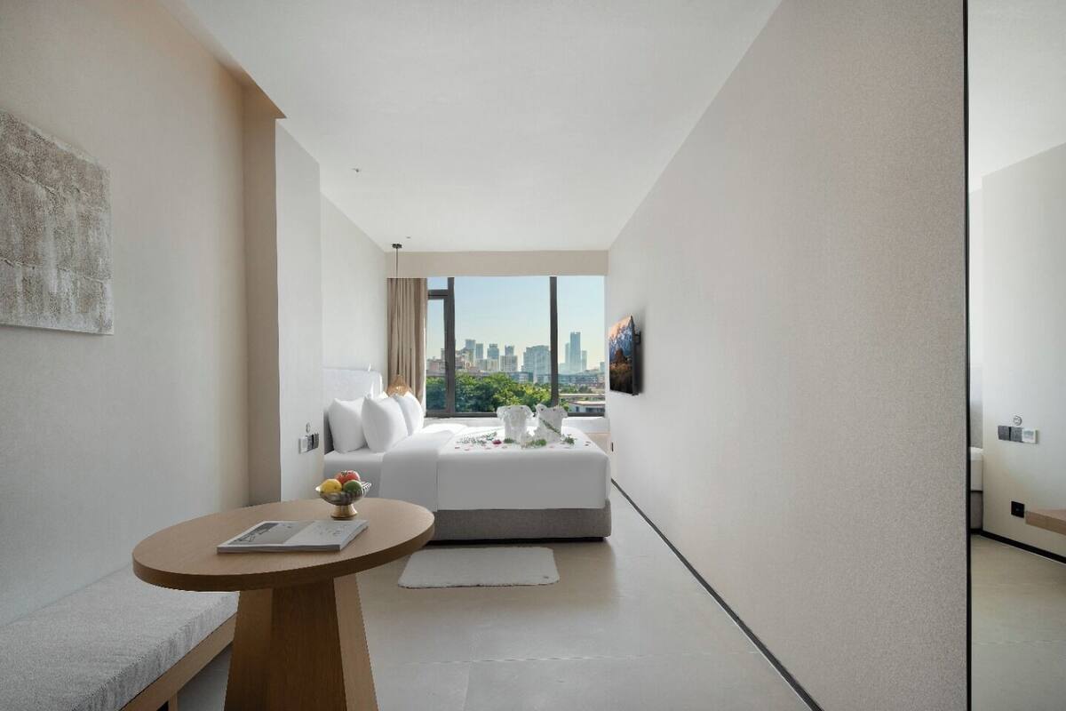 Una habitación de hotel moderna con vistas a la ciudad, una cama, una mesita con un libro y un jarrón con flores.
