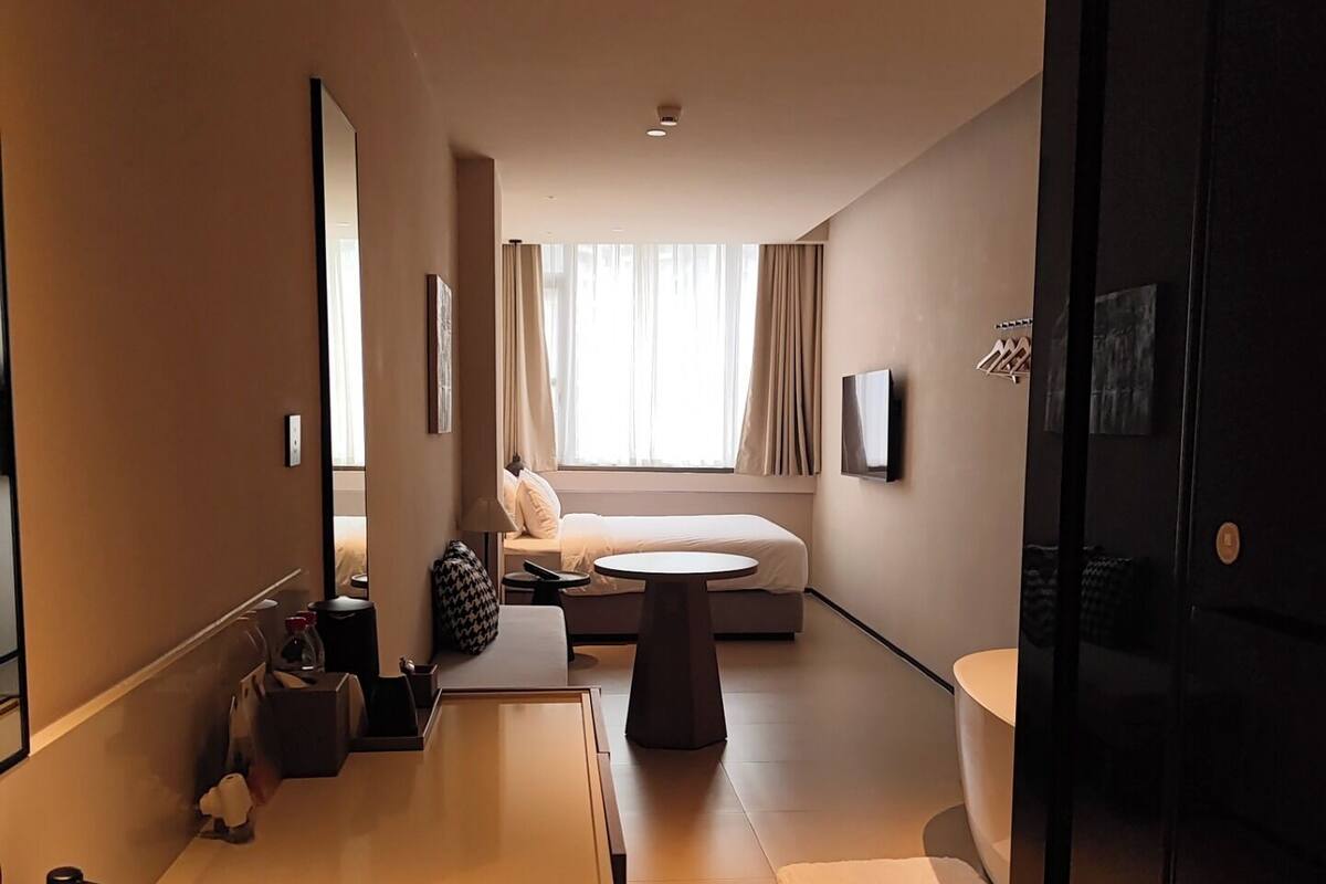 Una habitación de hotel moderna con una cama grande, una zona de estar, una mesita pequeña, una bañera y un espejo amplio.