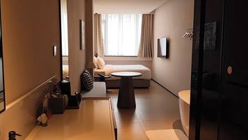 A modern hotel room with a large bed, a sitting area, a small table, a bathtub, and a large mirror.