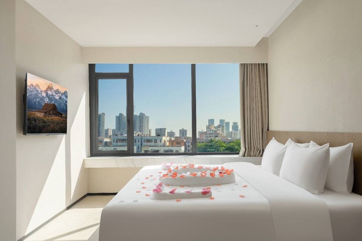 Habitación de hotel con un ventanal grande, cama con ropa de cama blanca, un cuadro en la pared y vistas a un paisaje urbano.